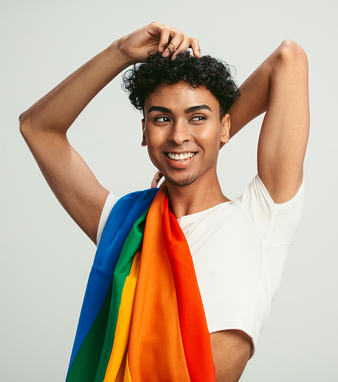 Smiling person with short black curly hair, standing with arms overhead, wearing a white shirt and draped with a rainbow pride flag.