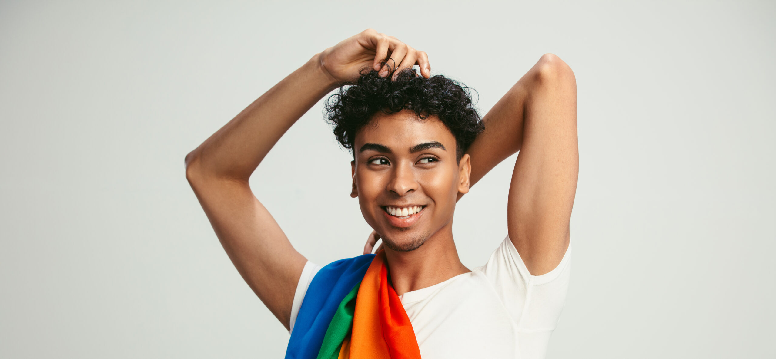 Smiling person with arms above head, curling black hair, weairing a white crop-top with a rainbow flag draped over their shoulder.