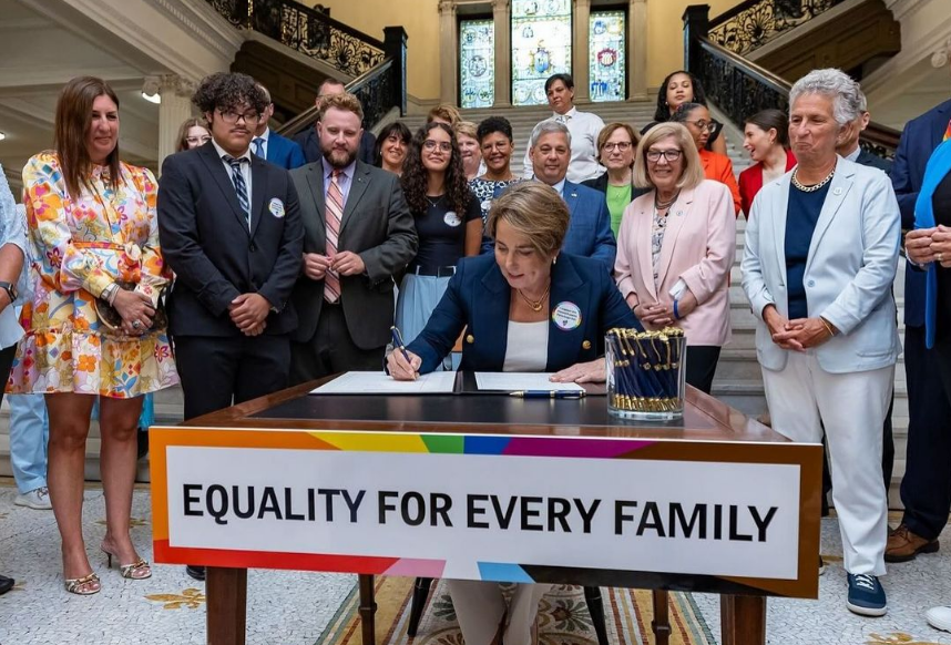 Smiling people standing around and behind a person sitting at a desk signing legislation. 