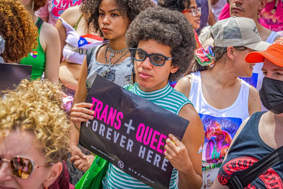 People in colorful clothes standing at a protest, one with a sign that says TRANS + QUEER forever here