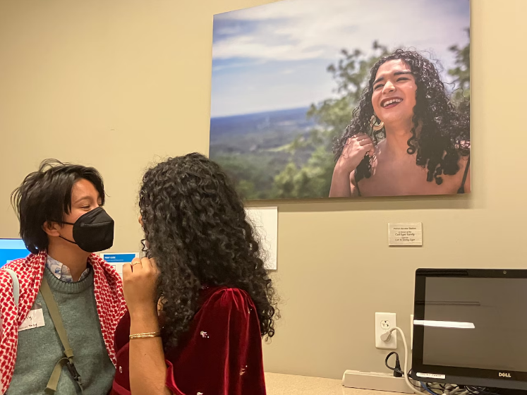 Two people, one with long black curly hair and the other wearing a black mask standing in front of a large photo of a person smiling outside near trees with long black curly hair.