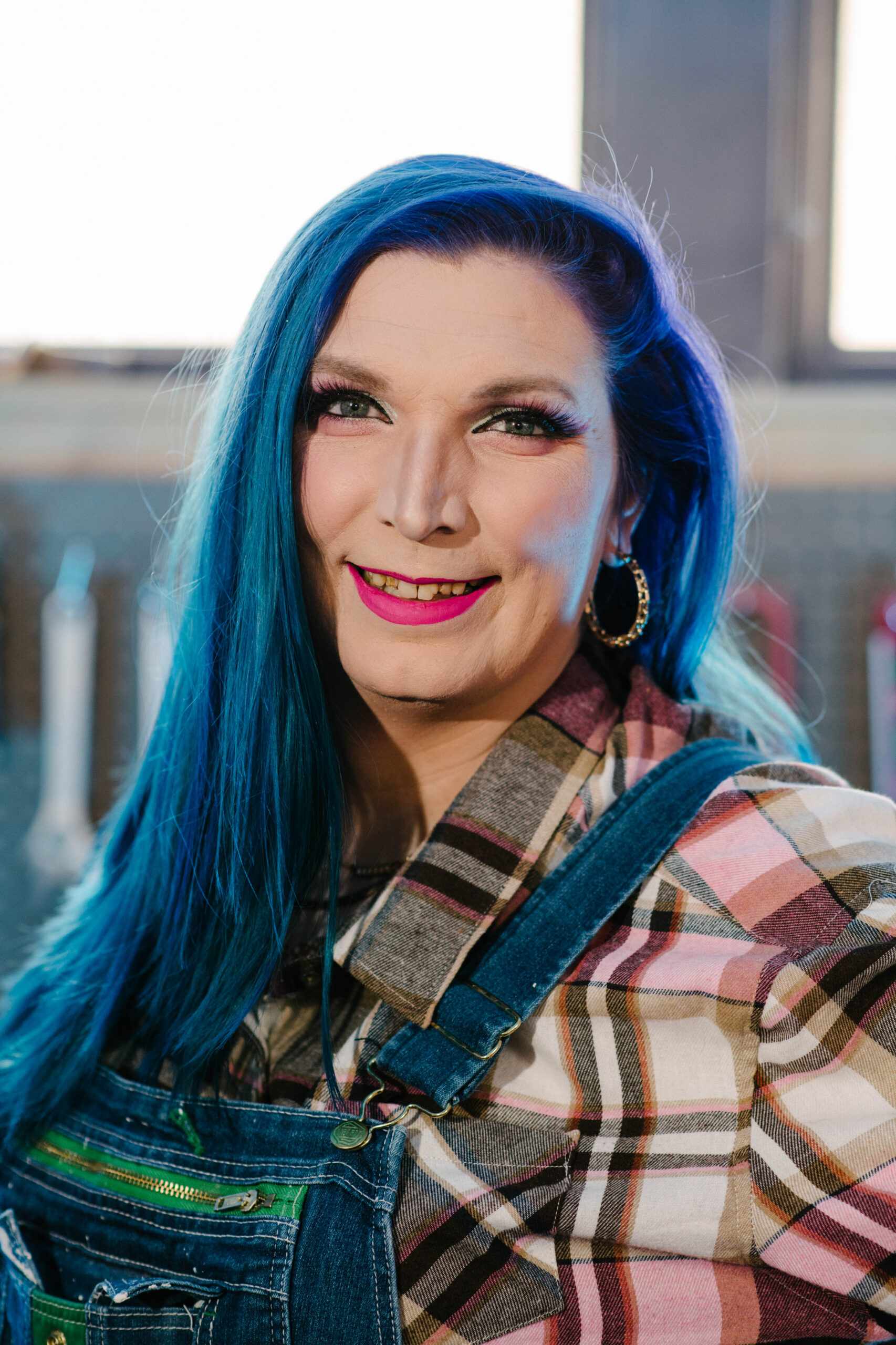 Headshot of Mercury Stardust smiling. She has long blue hair and is wearing denim overalls and a flannel shirt.